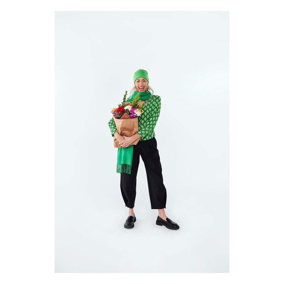 A cheerful woman smiles broadly, holding a paper bag filled with colorful flowers in a brightly lit studio setting, wearing green and black fashion.