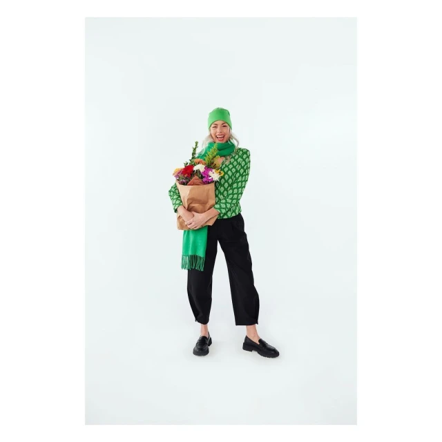 A cheerful woman smiles broadly, holding a paper bag filled with colorful flowers in a brightly lit studio setting, wearing green and black fashion.