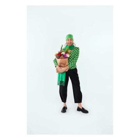 A cheerful woman smiles broadly, holding a paper bag filled with colorful flowers in a brightly lit studio setting, wearing green and black fashion.
