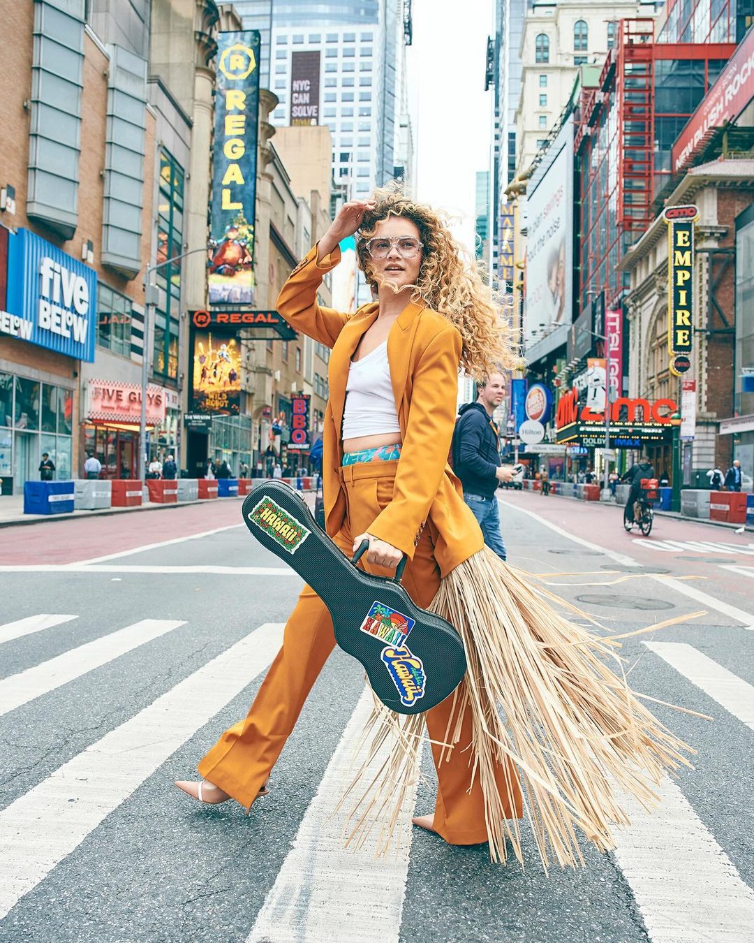 Woman in orange suit walks across crosswalk in New York City, holding ukulele case with straw fringe, wearing glasses and white top.
