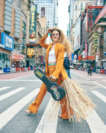 Woman in orange suit walks across crosswalk in New York City, holding ukulele case with straw fringe, wearing glasses and white top.