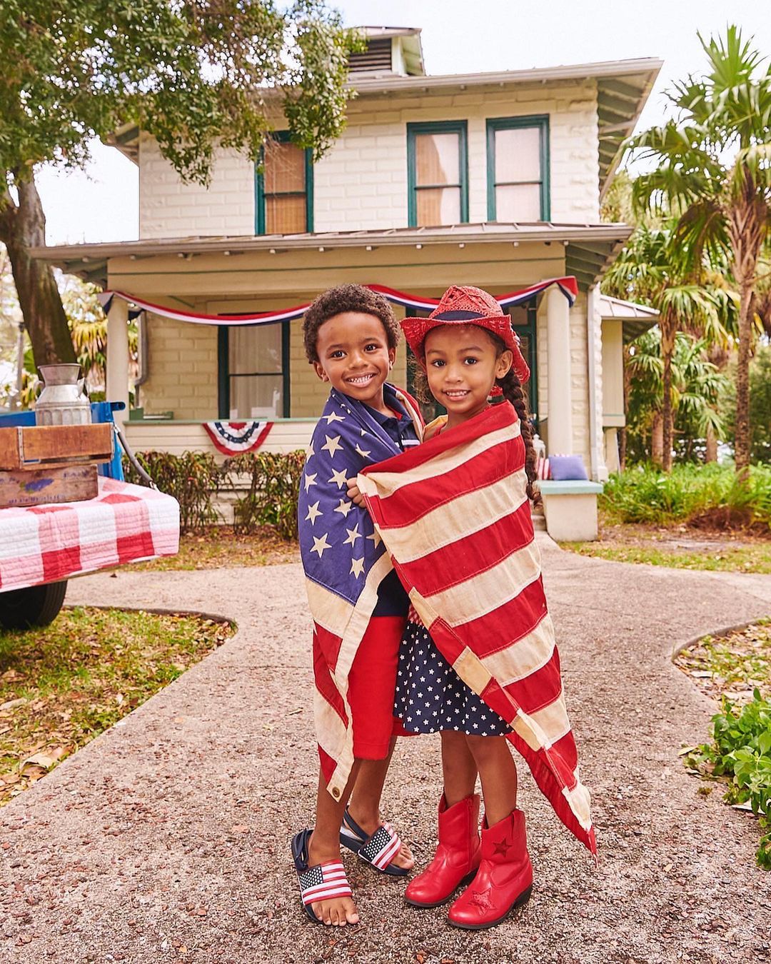 Two smiling children wrapped in the American flag in front of a two-story house decorated with patriotic bunting on a sunny day.