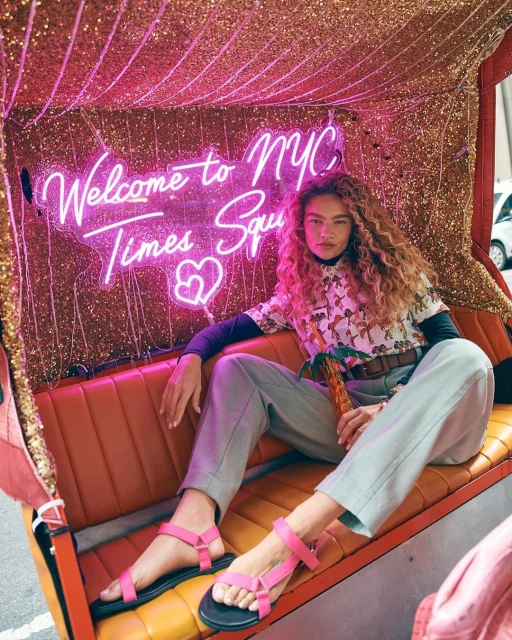 Woman with curly hair wearing a printed shirt and gray pants sits on a taxi with a neon sign reading "Welcome to NYC Times Square".