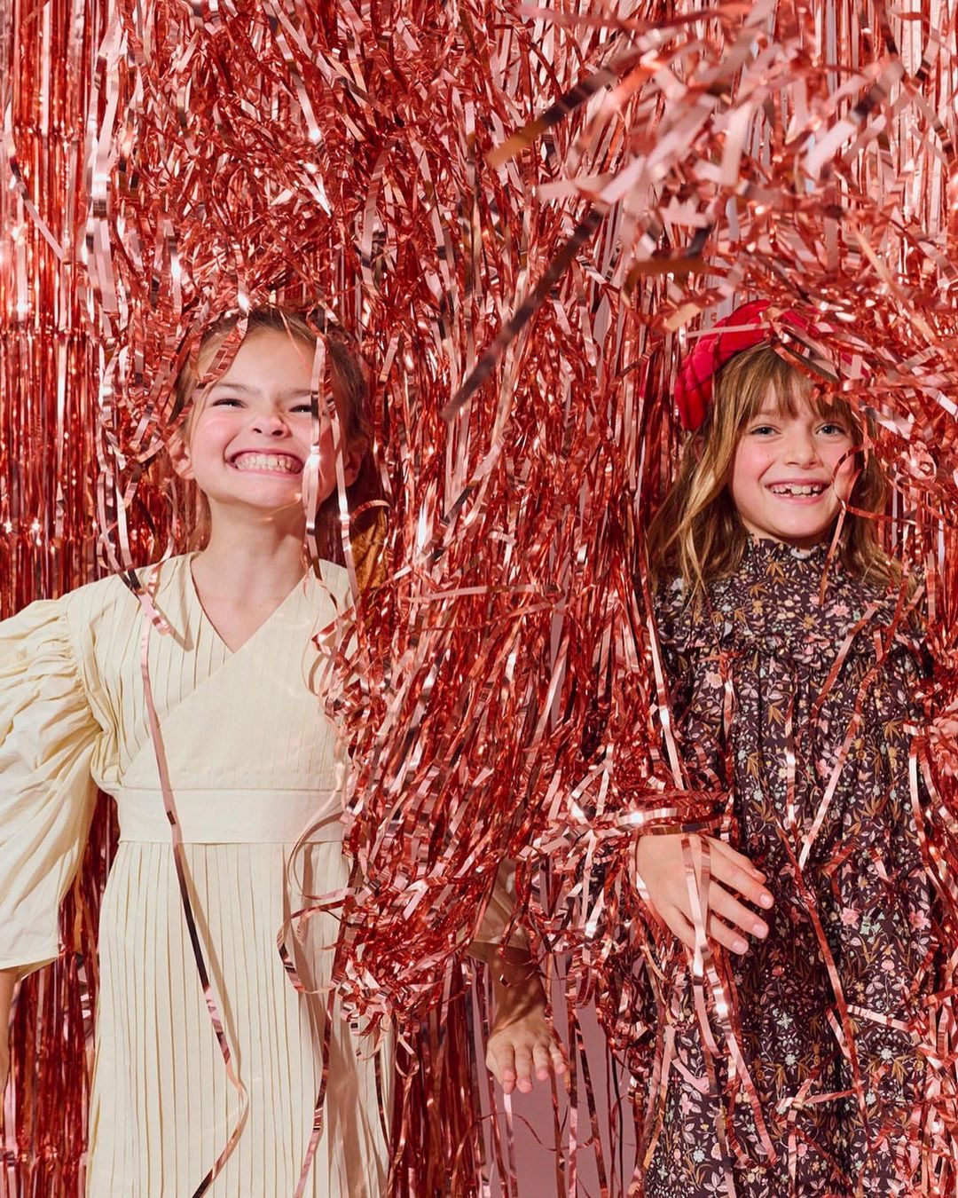 Two smiling girls with long hair pose among pink metallic streamers indoors. Both wear dresses and look at the camera.