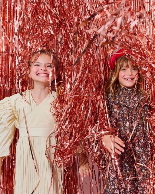 Two smiling girls with long hair pose among pink metallic streamers indoors. Both wear dresses and look at the camera. 
