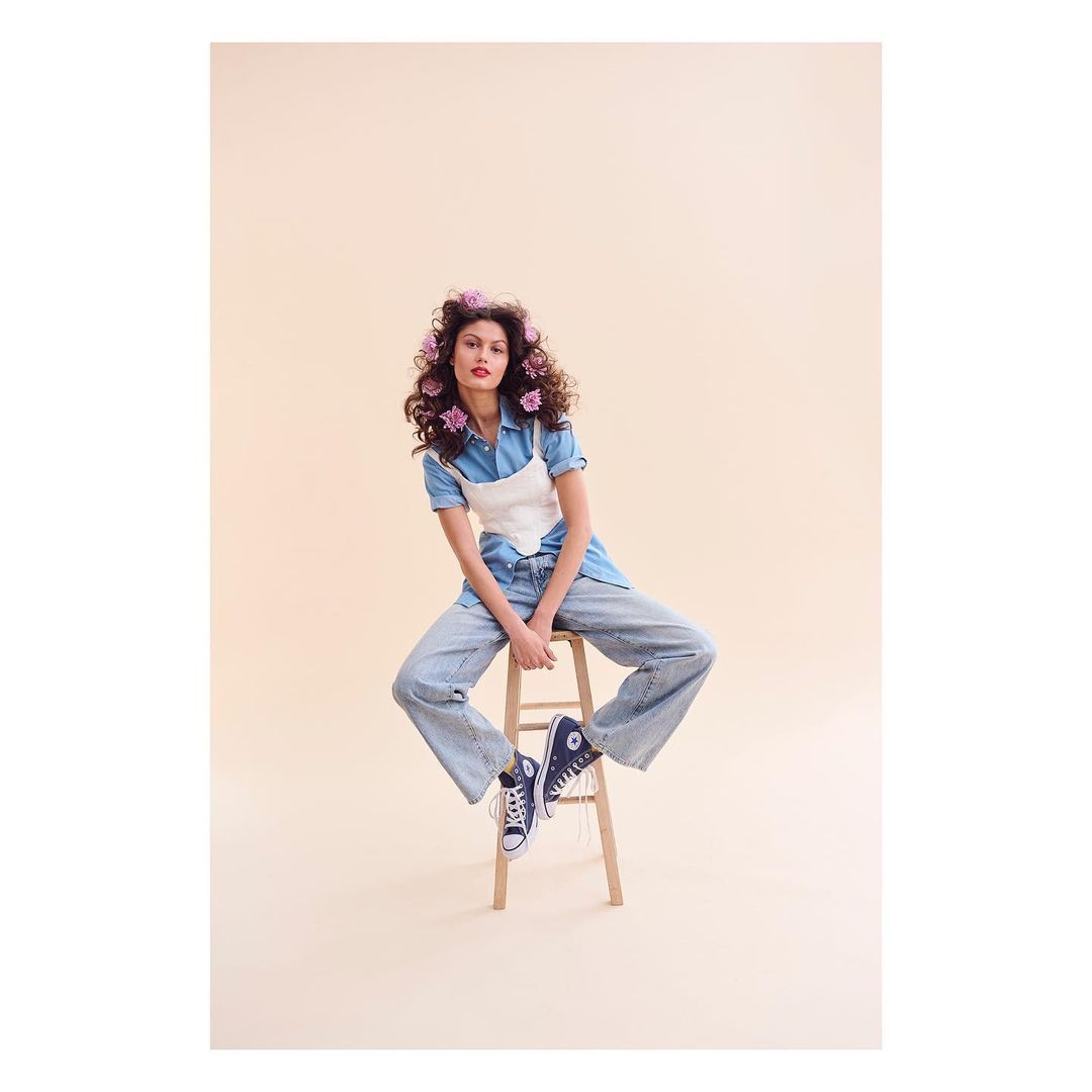 A young woman with curly hair and pink flowers sits on a wooden stool wearing denim and sneakers against a soft beige backdrop.