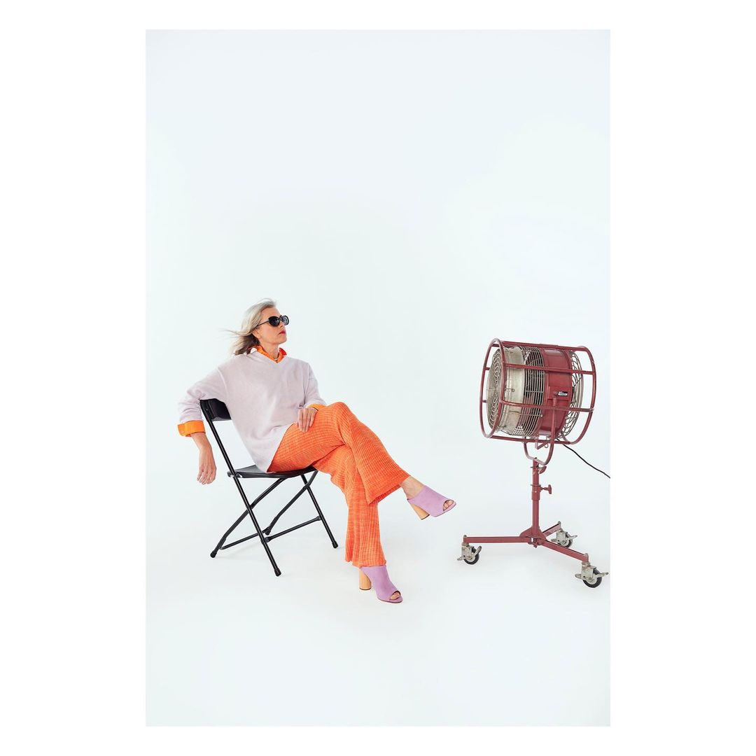 Woman in orange pants and sunglasses sits on a chair next to a red retro fan in a bright photo studio setting. Fashion photography.
