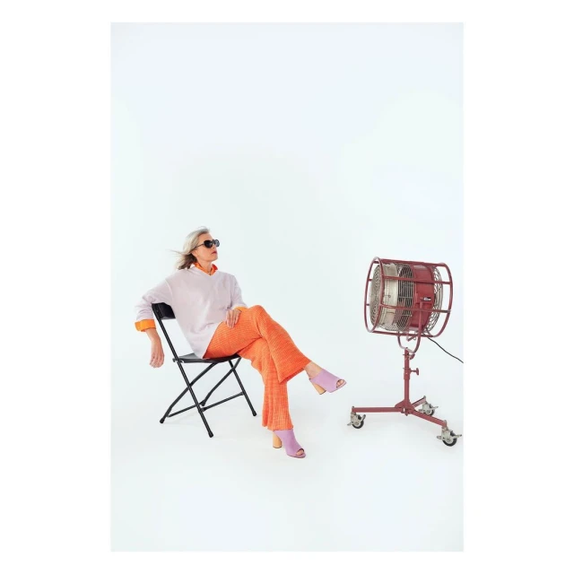 Woman in orange pants and sunglasses sits on a chair next to a red retro fan in a bright photo studio setting. Fashion photography.