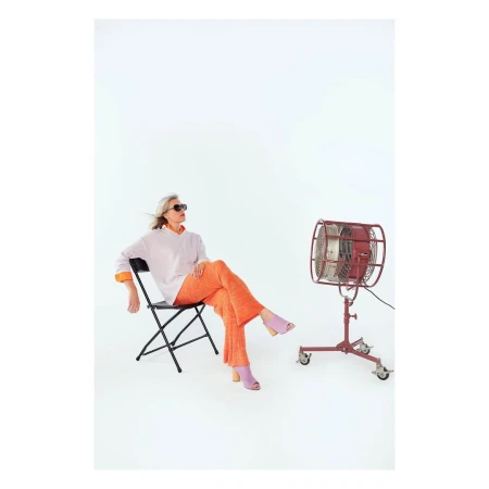 Woman in orange pants and sunglasses sits on a chair next to a red retro fan in a bright photo studio setting. Fashion photography.