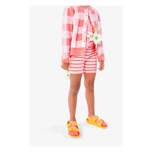 Child stands in a studio setting, wearing a pink gingham cardigan, pink striped shorts and top, and yellow sandals holding a daisy.