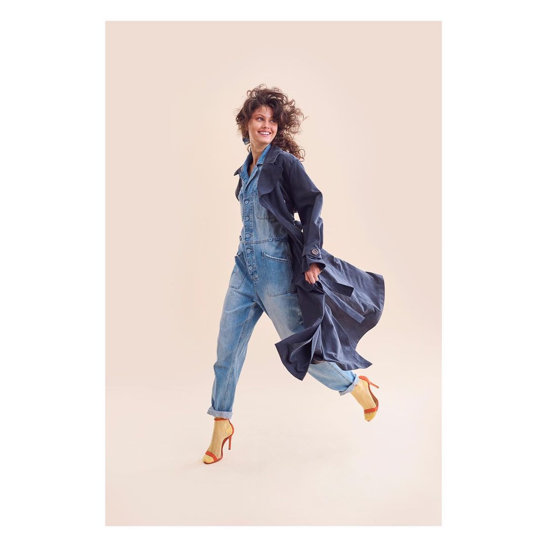A woman with curly hair smiles and leaps in a denim jumpsuit and coat, accessorized with heeled sandals, against a light peach backdrop.