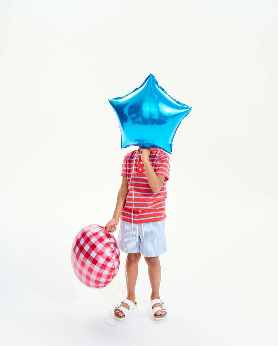 Child stands in studio holding a red-and-white checkered balloon and a blue star balloon covering their face, against a white backdrop.