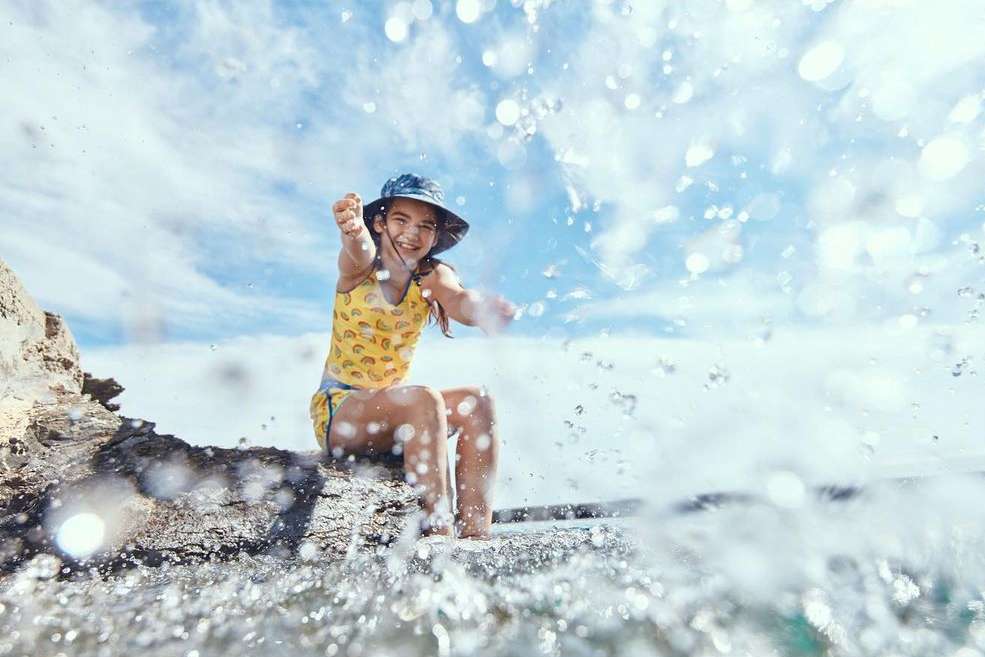 A young child smiles while seated on a rock formation at the beach, with water splashes in foreground, wearing a yellow bathing suit and bucket hat.