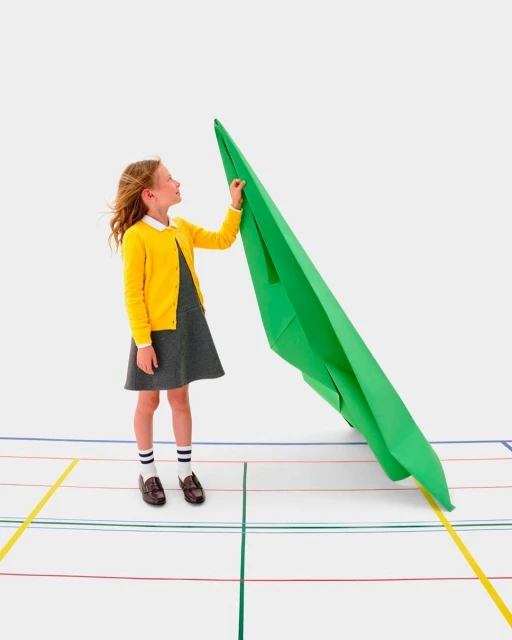 A young girl in school uniform stands next to a giant green paper airplane on a white surface with colored lines, creating a playful and imaginative scene.