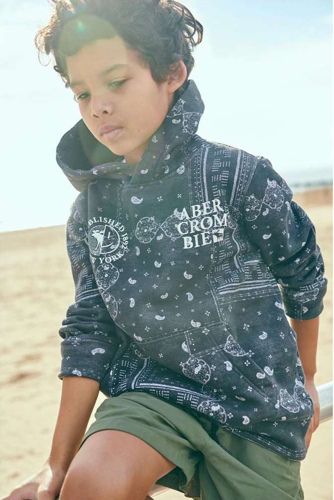 Child models Abercrombie & Fitch hooded sweatshirt outdoors. The boy sits smiling in natural daylight in front of a blurred background.