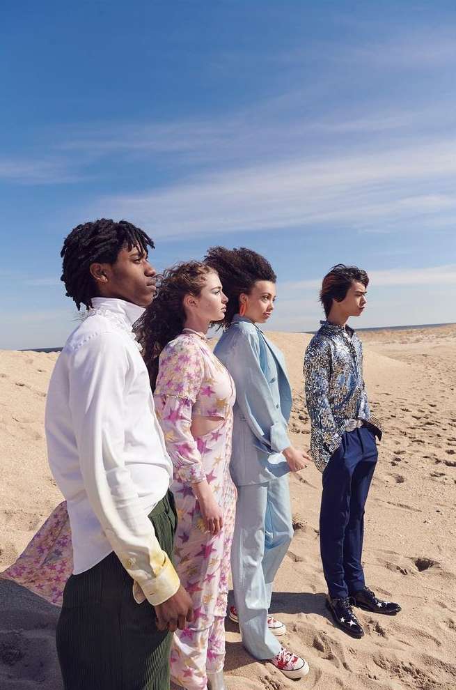 Group of four diverse young adults standing together on a sandy beach under a sunny blue sky, wearing stylish pastel-colored clothing.