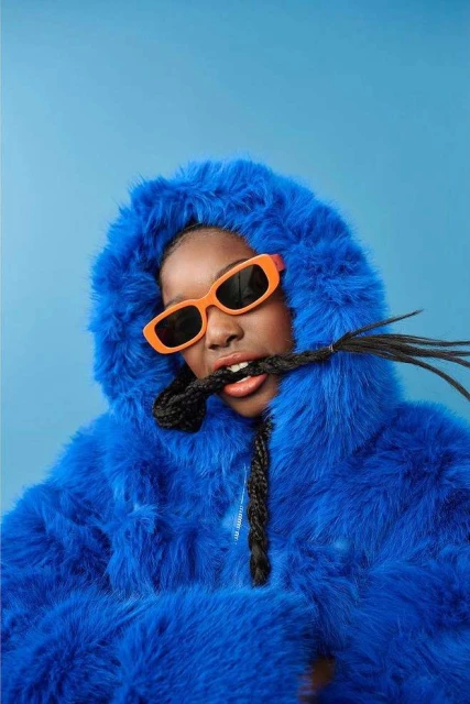Portrait of a black female wearing orange sunglasses and a blue hooded coat biting a braid against a solid blue studio backdrop.