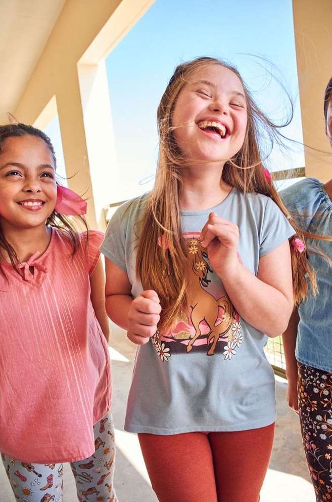 Three smiling girls, including a child with Down syndrome, pose in front of a white colonnade outdoors on a sunny day.