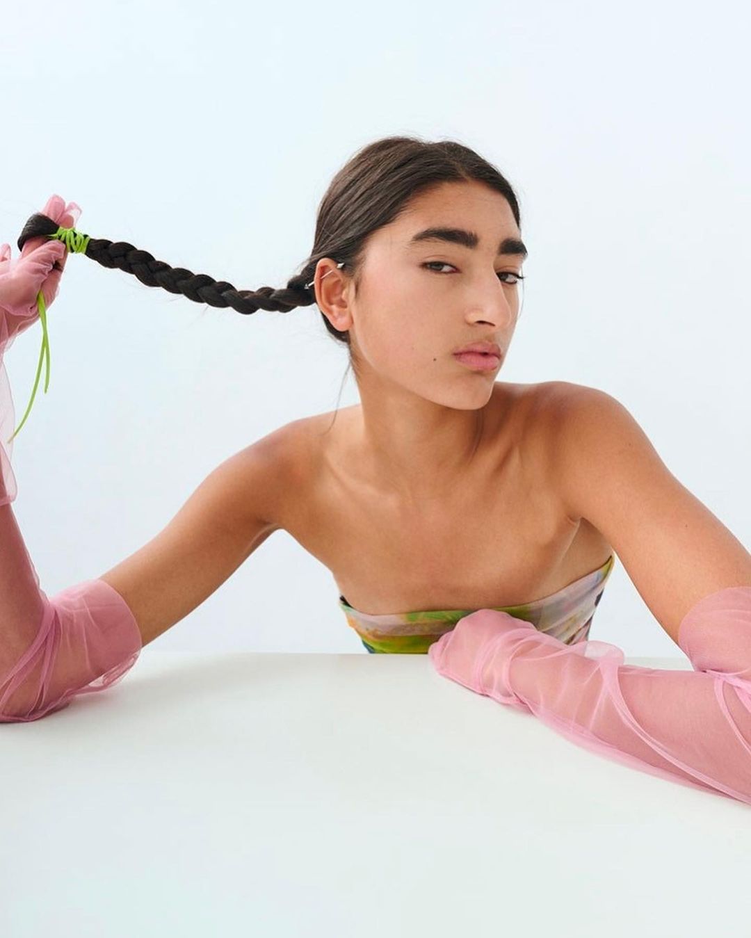 Woman poses in studio wearing pink gloves and a patterned top, holding a braided plait adorned with green string against a white backdrop.