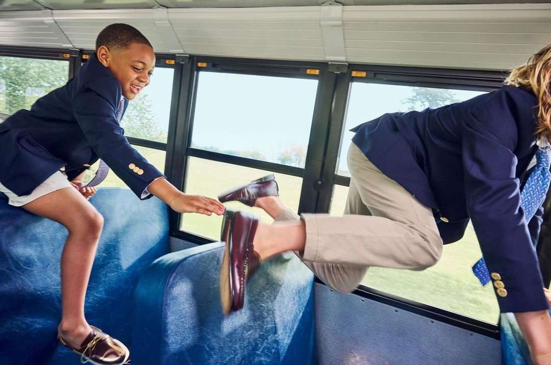 Two children in school uniforms climbing over bus seats, one reaching out a hand, creating a sense of fun and camaraderie.