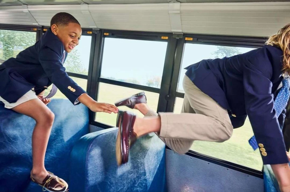 Two children in school uniforms climbing over bus seats, one reaching out a hand, creating a sense of fun and camaraderie.