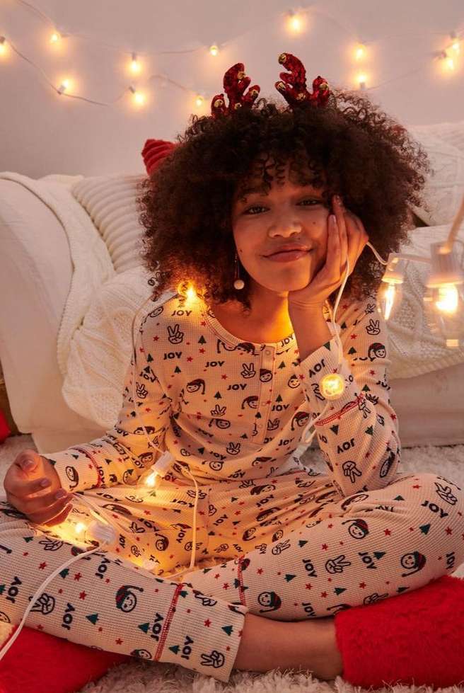 A young adult female with curly hair sits cross-legged in patterned pajamas with holiday reindeer antlers, surrounded by string lights.