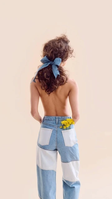 Back view of a slender adult woman with curly hair, wearing denim pants and a blue bow, with yellow flowers in the back pocket.