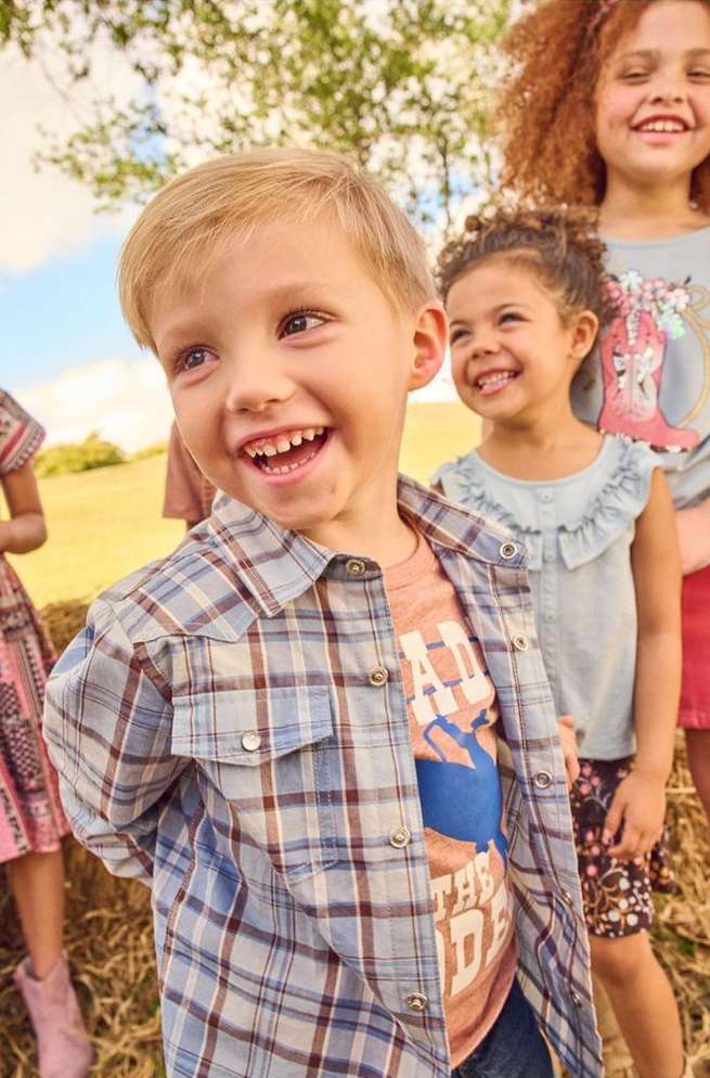 Smiling boy in a plaid shirt with two smiling girls standing behind him in a grassy outdoor setting on a sunny day.