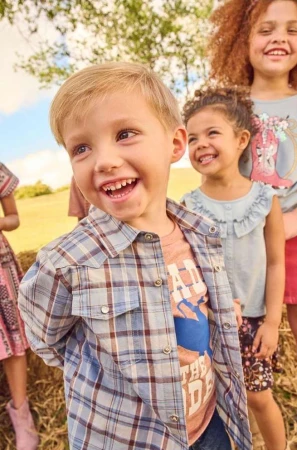 Smiling boy in a plaid shirt with two smiling girls standing behind him in a grassy outdoor setting on a sunny day.