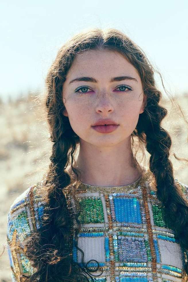 Close-up portrait of a young woman with freckles and braided hair, wearing a beaded top against a bright, out-of-focus background.