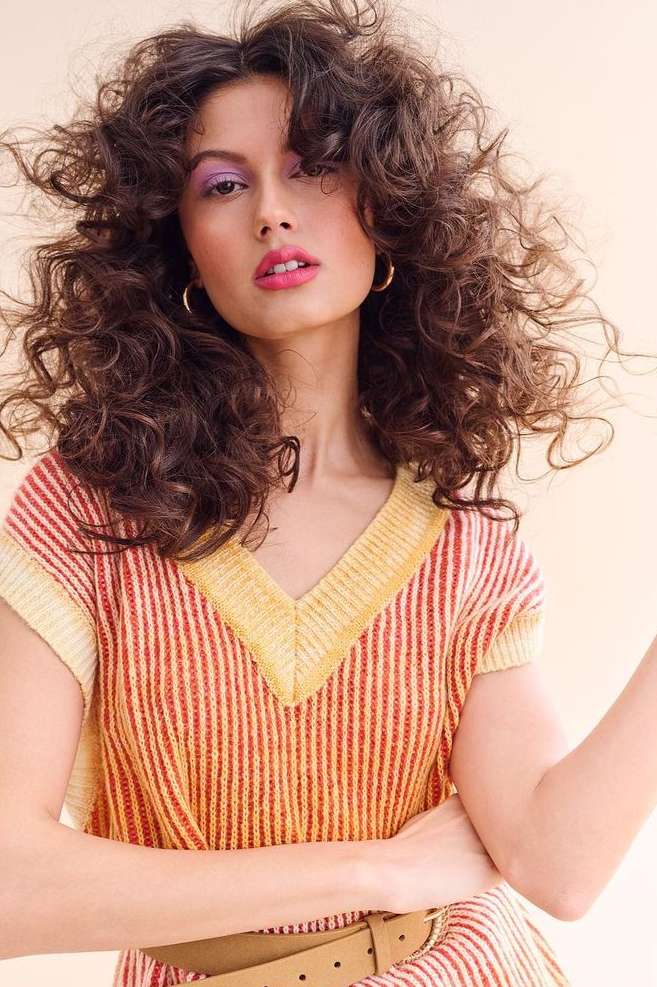 Brunette adult woman with curly hair wearing a striped top poses in a studio shot with neutral background and hoop earrings.