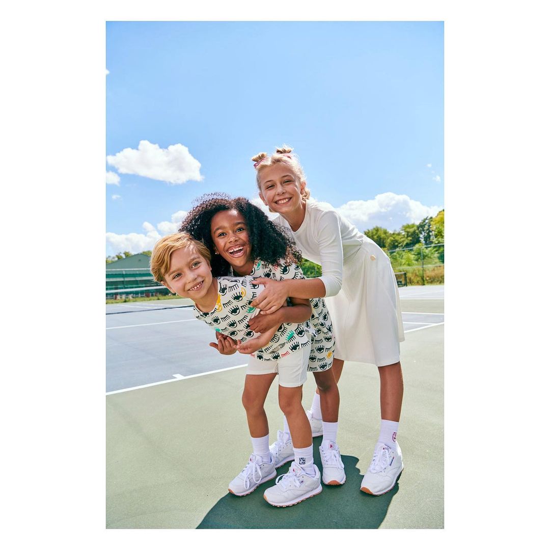 Three smiling children outdoors: a boy and girl in matching eye-print outfits, and a taller girl in a white dress stand together.