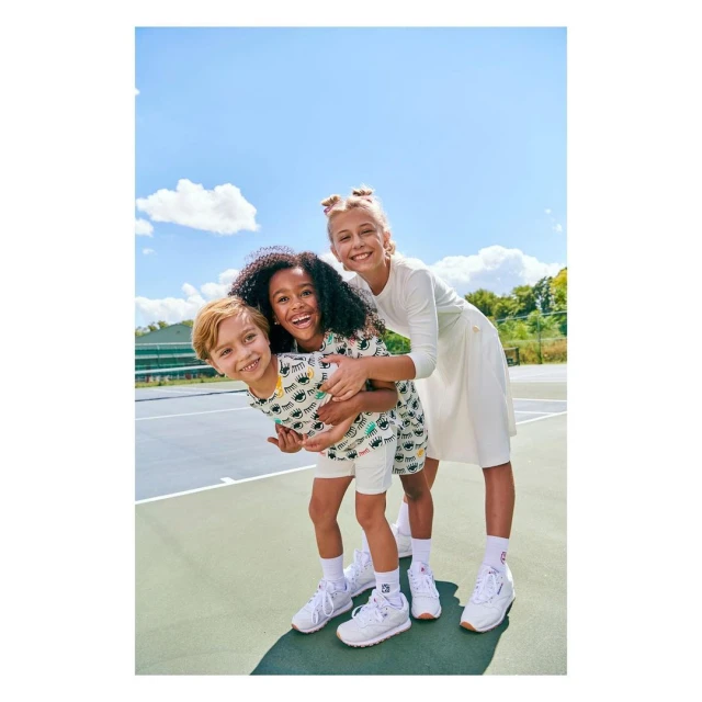 Three smiling children outdoors: a boy and girl in matching eye-print outfits, and a taller girl in a white dress stand together.
