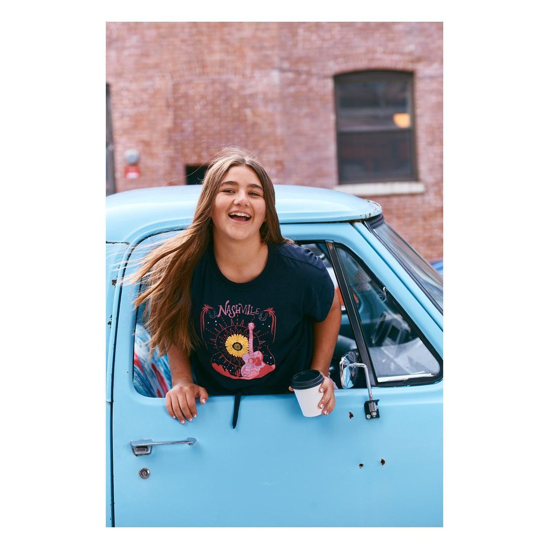 Smiling young woman with long brown hair leans out the window of a light blue truck while holding a disposable coffee cup.