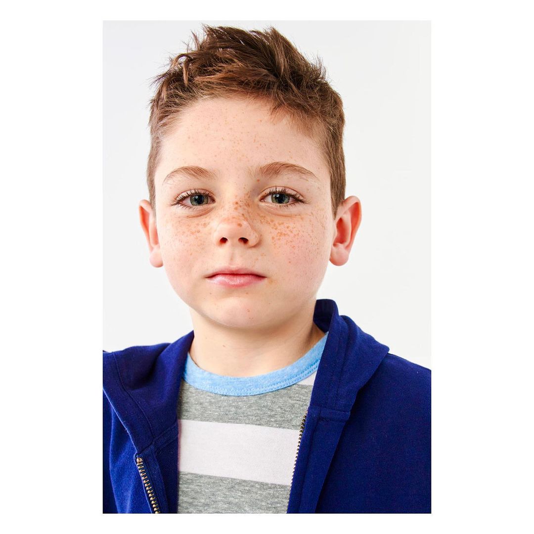 Close-up studio portrait of a young boy with freckles wearing a blue zip-up hoodie and striped t-shirt against a white background.