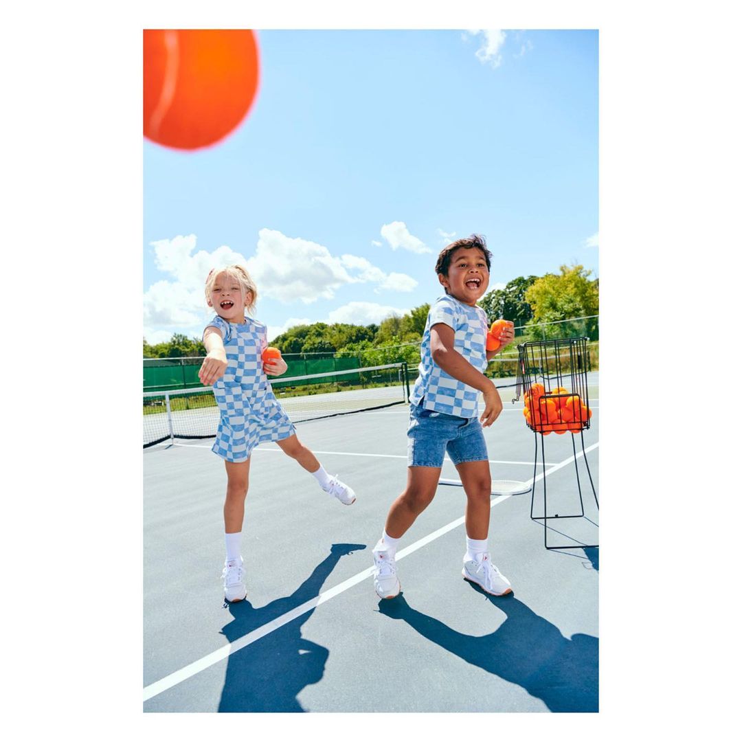 Two children, a boy and a girl, play tennis on an outdoor court on a sunny day. They are wearing matching blue checkerboard outfits.