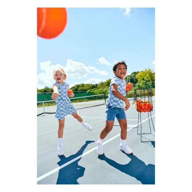 Two children, a boy and a girl, play tennis on an outdoor court on a sunny day. They are wearing matching blue checkerboard outfits.