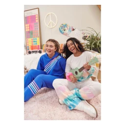 Two smiling teenage girls seated on a fluffy pink carpet playing a small light green ukulele in a brightly decorated bedroom.