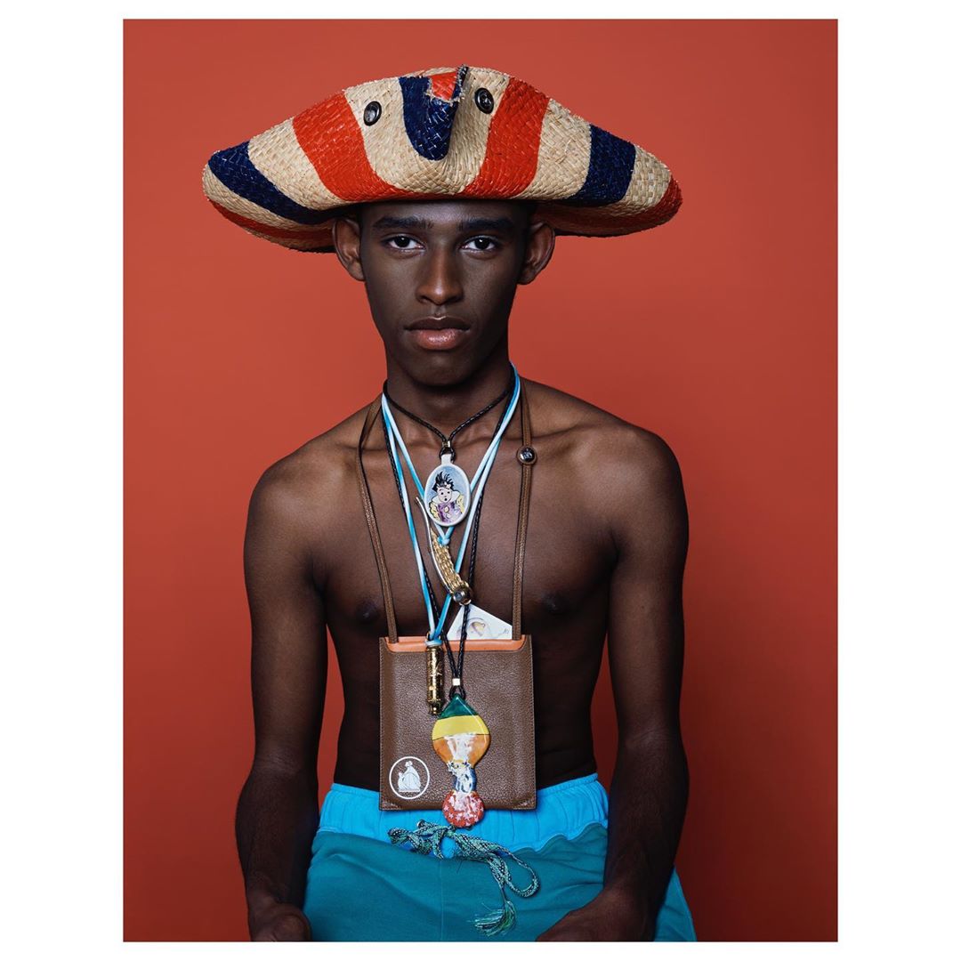 Young Black male in an orange studio background wearing a blue and red hat and a variety of beaded necklaces and a purse.