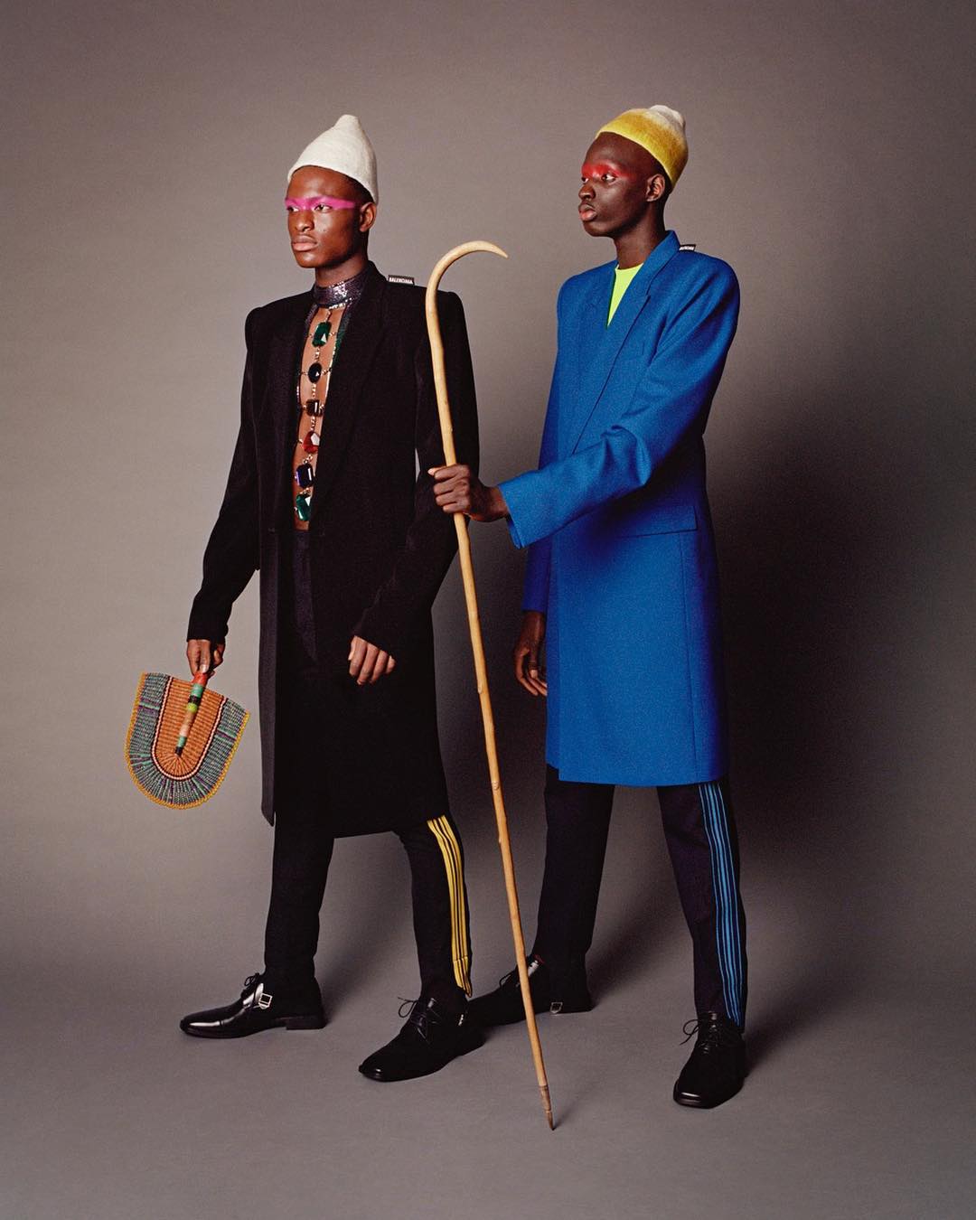 Two Black men in elegant blazers, one blue, one black, stand in a studio against a gray background, one holding a cane and a fan.