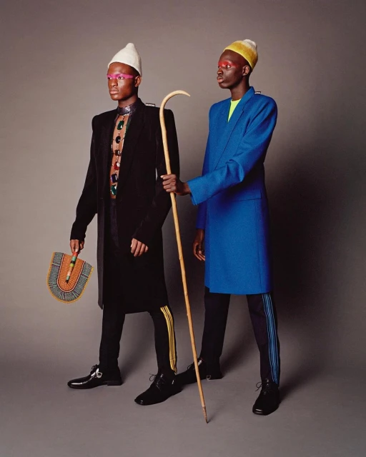 Two Black men in elegant blazers, one blue, one black, stand in a studio against a gray background, one holding a cane and a fan.