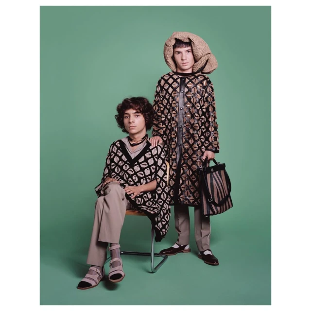 Two young male fashion models pose against a green backdrop, wearing patterned outerwear, tan trousers, and sandals; one holds a tote bag.