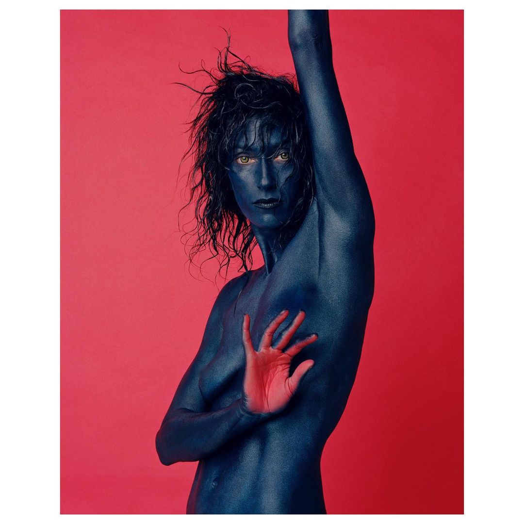 Adult with blue body paint posing with hand, set against a pink background in a studio-style portrait with dramatic visual appeal.