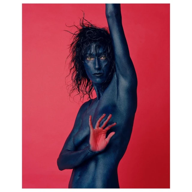 Adult with blue body paint posing with hand, set against a pink background in a studio-style portrait with dramatic visual appeal.