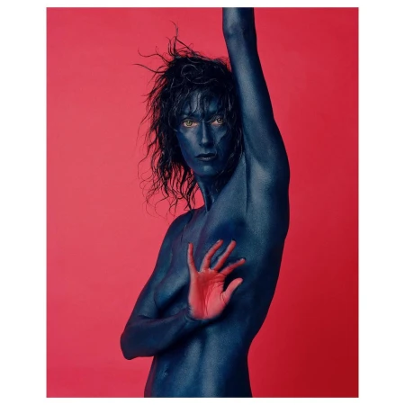 Adult with blue body paint posing with hand, set against a pink background in a studio-style portrait with dramatic visual appeal.