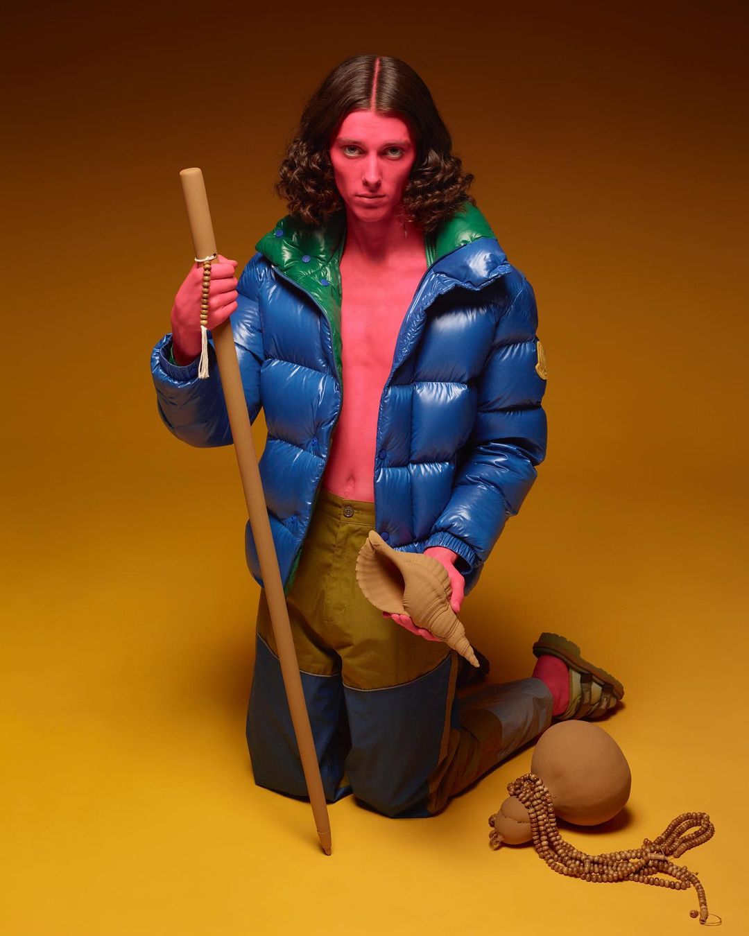 A person with red skin and long brown hair poses kneeling in a studio, wearing a blue jacket, holding a staff and shell, against a yellow backdrop.