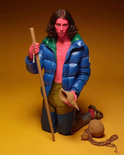 A person with red skin and long brown hair poses kneeling in a studio, wearing a blue jacket, holding a staff and shell, against a yellow backdrop.