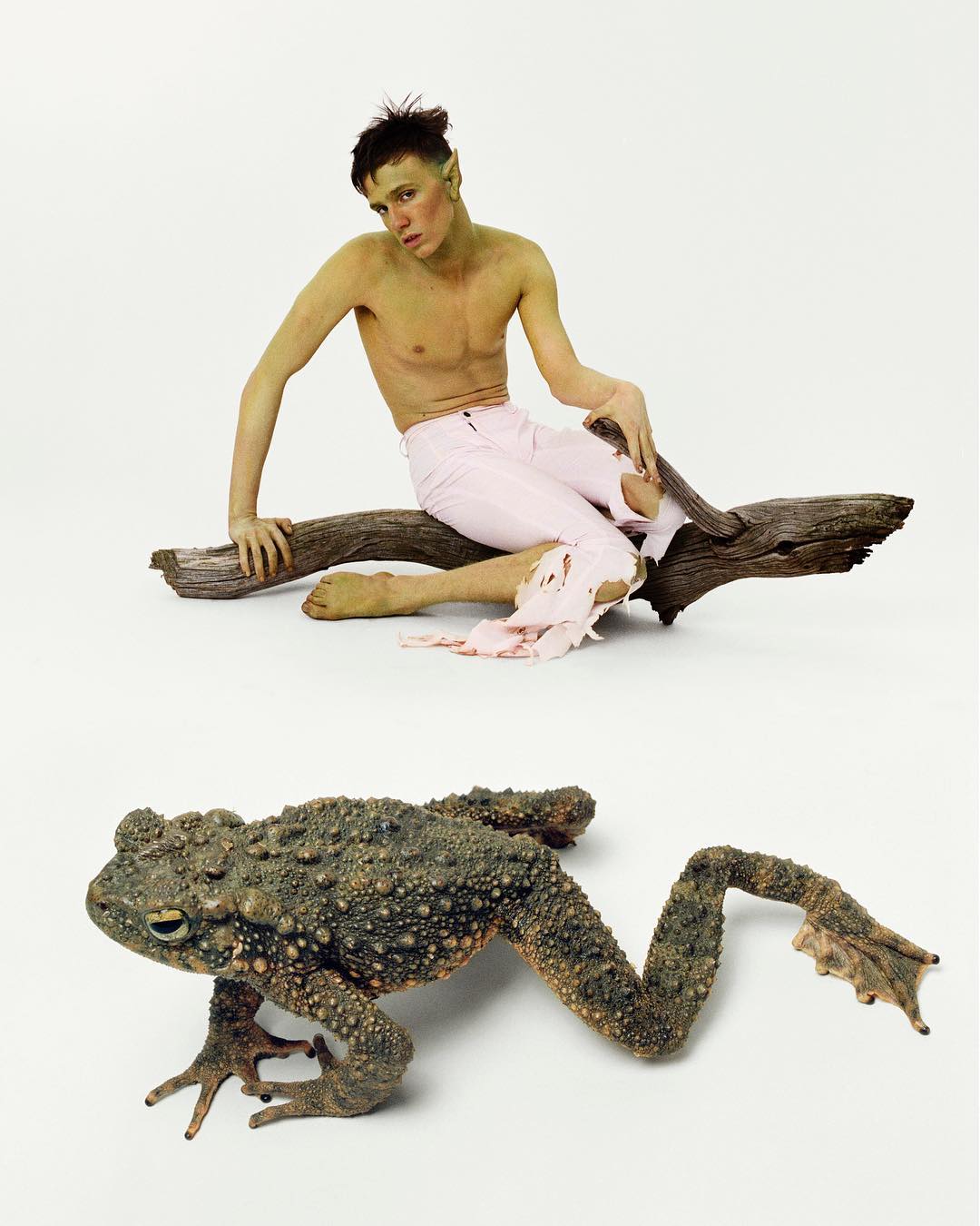 Photo of a shirtless male model with green skin sitting on a log, next to a large frog with bumpy skin, against a white backdrop.