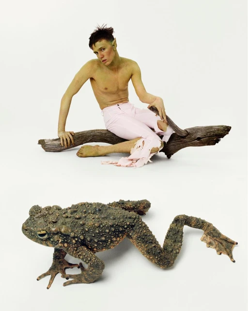 Photo of a shirtless male model with green skin sitting on a log, next to a large frog with bumpy skin, against a white backdrop.