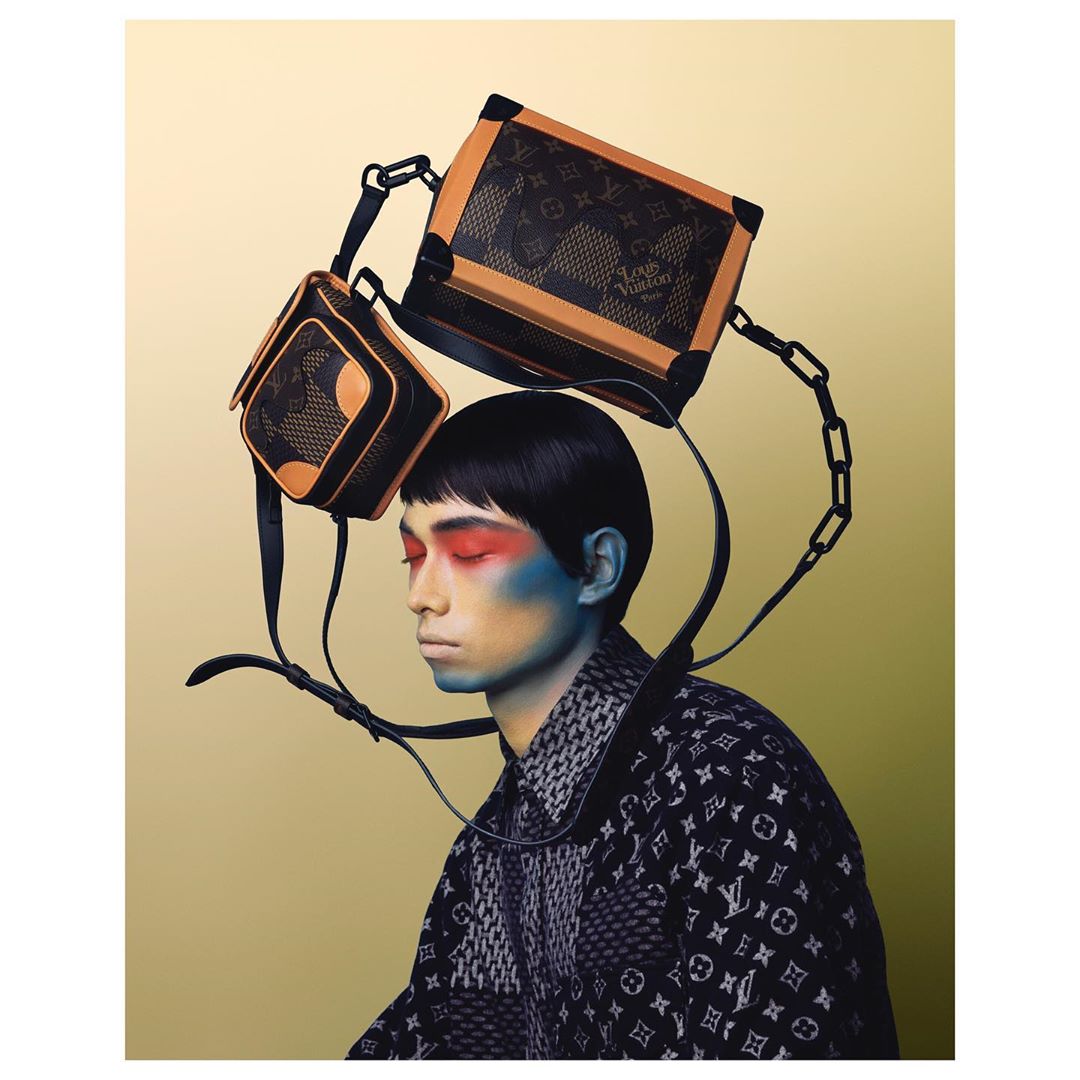 Editorial shot of a person with red and blue makeup, wearing a patterned jacket, and two black and tan bags floating above the head.